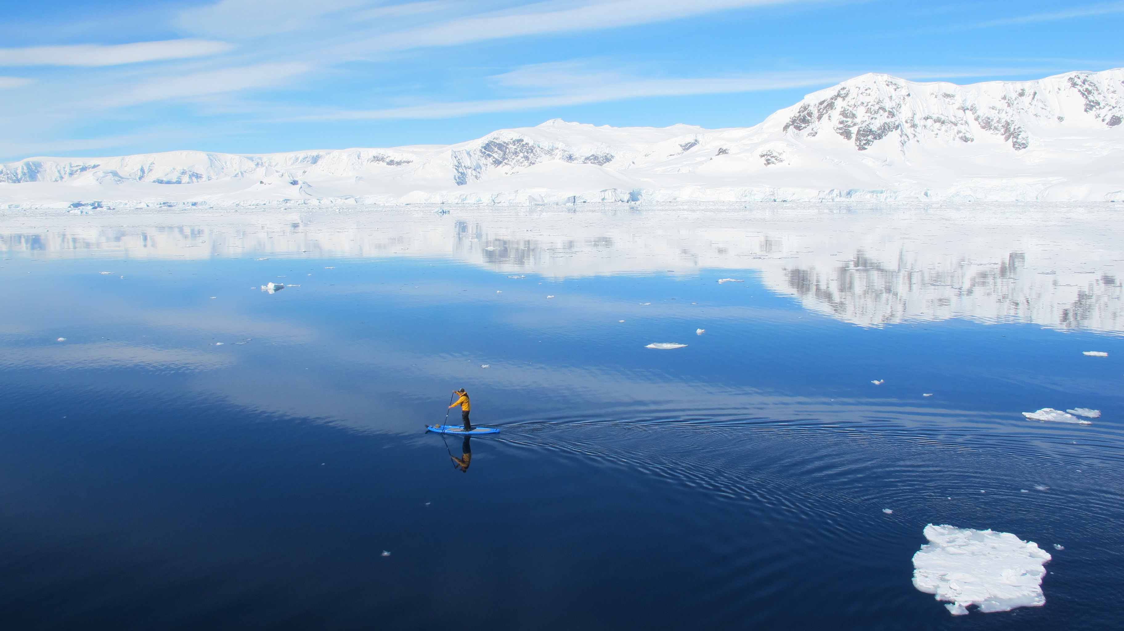 A passenger glides over the surface of pristine, glass-still Antarctic waters on a standup paddleboard.